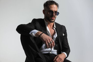 portrait of attractive bearded man with undone bowtie and shirt, wearing black tuxedo and sunglasses, holding knee up and posing in a fashion manner clipart