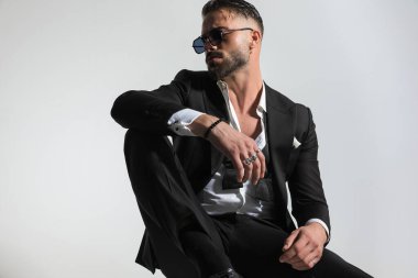 Stüdyoda gri bir arka planda otururken dizini kaldırıp yan yan yan yan yan bakan dağınık papyonlu şık bir adam.