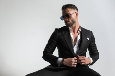 Siyah smokini ayarlarken ve düğmelerini iliklerken yana doğru bakan açık yakalı sakallı genç adamın portresi gri arka planda stüdyoda oturuyor.