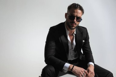 Siyah smokinli seksi damadın portresi moda pozunda oturuyor ve stüdyoda gri arka planda havalı duruyor.