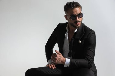 Siyah smokinli baştan çıkarıcı genç adam dizinin üzerinde dirseğini tutuyor, gözlerini kaçırıyor ve stüdyoda gri arka planda poz veriyor.