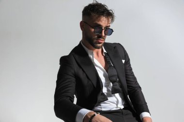 Havalı, tıraşsız, yakası açık bir adam stüdyodaki gri arkaplanın önüne bakıyor.