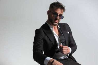 Güneş gözlüklü yakışıklı adam, siyah smokin giyiyor. Stüdyoda, gri arka planda, kendine güvenen bir şekilde poz veriyor.