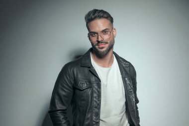 Siyah deri ceketli ve gözlüklü gri arka planda poz veren mutlu bir adam.