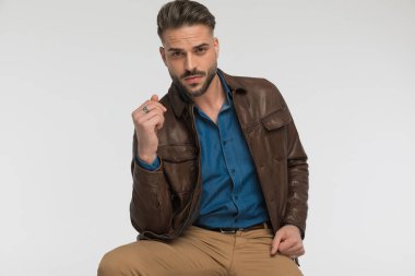 Kahverengi deri ceketli seksi bir adam moda kollarıyla kollarını tutuyor ve açık gri arka planda otururken poz veriyor.