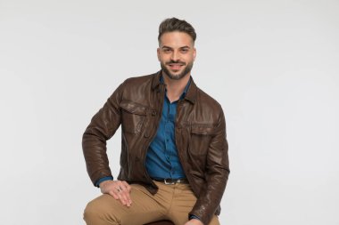 Kahverengi ceketli yakışıklı genç adam gri arka planda stüdyoda gülümsüyor ve oturuyor.