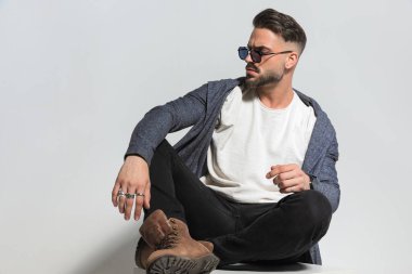 Kapüşonlu, kapüşonlu, güneş gözlüklü, dirseğini dizine dayayıp gri arka planda poz veren havalı adam.