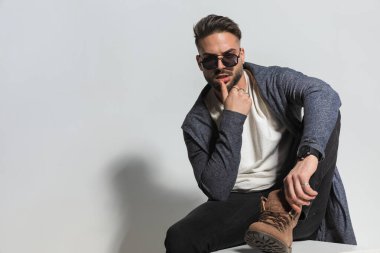 Kapüşonlu şehvetli genç adam bir eli baştan çıkarıcı bir şekilde dudaklara dokunuyor, diğer dirseğini dizinin üstüne koyup gri arka planda poz veriyor.