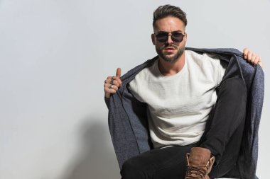 Stüdyoda gri arka planda kendine güvenen güneş gözlüklü genç adam kapüşonluyu çekiyor ve ayarlıyor.