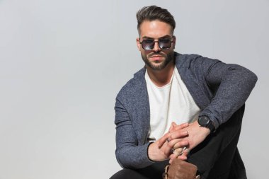 Gözlüklü, el ele tutuşan ve gri arka planda dirseğiyle poz veren kibirli sıradan bir adam.