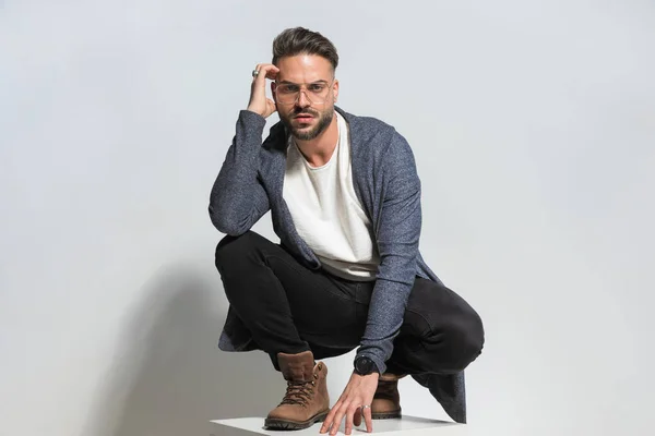sexy bearded man in cool outfit holding arms in fashion pose, touching head and thinking, holding elbow on knee and posing in studio