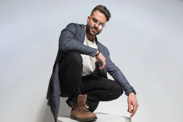 unshaved guy with beard and glasses holding elbow on knee and crouching, posing in a cool way in studio