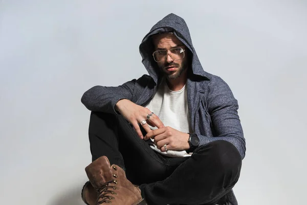 mysterious seated man with hoodie on head holding elbows on knees, touching fingers and posing with bend knee in studio on grey background