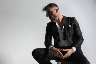 Smokinli havalı sakallı adam, yakası açık, parmaklarına dokunuyor, gri arka planda şık bir poz veriyor.