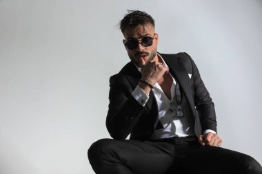 Smokinli, yakası açık genç bir damat stüdyodaki gri arka plan önünde dudaklarına dokunuyor.