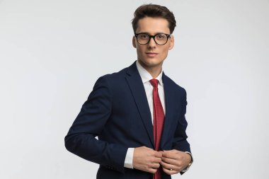 Gözlüklü mutlu genç işadamının portresi lacivert takımını ilikliyor ve stüdyoda gri arkaplan önünde gülümsüyor.