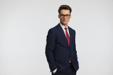 Lacivert takımlı çekici genç adam ceplerinde el ele tutuşuyor ve stüdyoda gri arka planda poz veriyor.