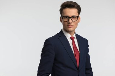 Lacivert takım elbiseli, kırmızı kravatlı, gözlüklü ve gri arka planda gülümseyen yakışıklı bir adamın portresi.