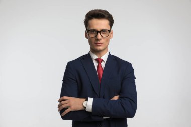 Şık takım elbiseli mutlu yakışıklı bir adamın portresi kollarını kavuşturup stüdyoda gri arka planda poz veriyor.