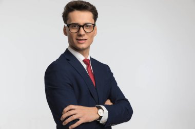 Mutlu genç işadamı lacivert takım elbise giyiyor kollarını kavuşturuyor ve stüdyoda gri arka planda gülümsüyor.