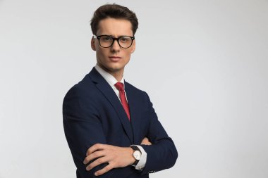 elegant young man with eyeglasses folding arms and posing in front of grey background in studio clipart