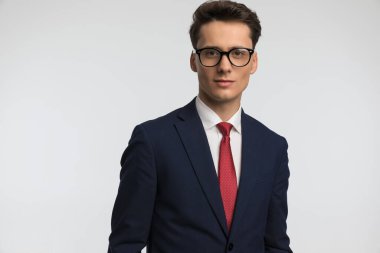 Gözlüklü mutlu bir adamın portresi kırmızı kravat takıyor ve stüdyoda gri arka planda gülümsüyor.