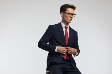 Kırmızı kravatlı, gözlüklü, takım elbiseli, stüdyoda gri arka planda oturan seksi bir genç.