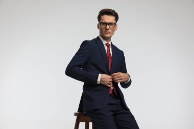 Zarif bir gencin portresi lacivert takımı tamir edip açıyor ve stüdyoda gri arka planda oturuyor.