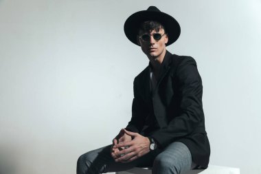 Güneş gözlüklü ve şapkalı yakışıklı adam stüdyoda gri arka planda otururken parmaklarına dokunuyor.