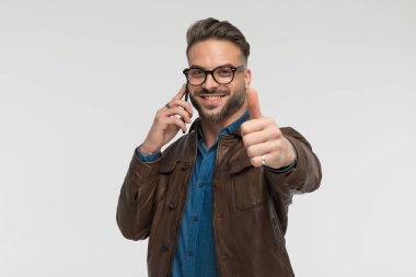 Mutlu adam telefonda konuşuyor, gülümsüyor ve stüdyoda gri arkaplan önünde baş parmak hareketi yapıyor.