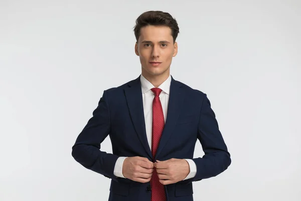 スタジオでグレーの背景の前でネイビーブルーのスーツを調整しながら笑顔で赤いネクタイを持つ誇り高いビジネスマン — ストック写真