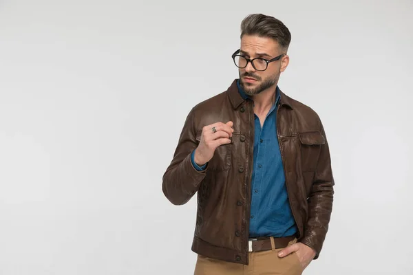 年轻帅气的男子 身穿棕色皮夹克 戴着眼镜 一边站在一边 一边一只手插在口袋里 一边站在工作室的灰色背景前 — 图库照片
