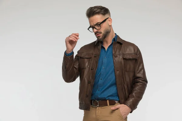attractive man with one hand in pocket looking down and posing while wearing blue denim shirt with brown leather jacket in front of grey background