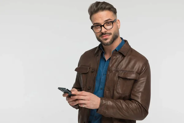 性感的留着胡子的男人穿着斜纹棉布衬衫 穿着棕色皮夹克 手持手机 写电子邮件 站在灰色背景前摆出姿势 — 图库照片
