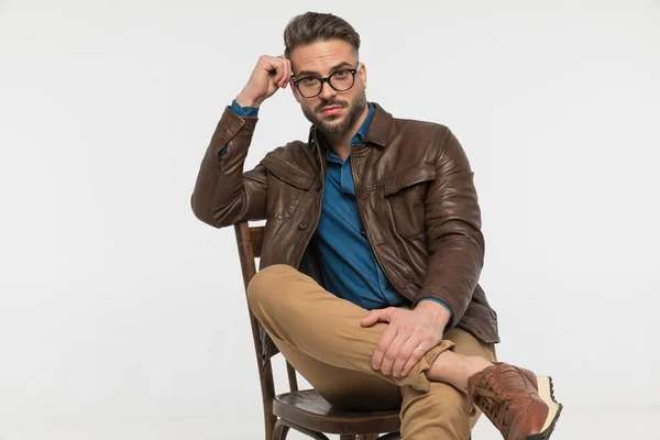 sexy thoughtful man in brown leather jacket, holding hand to temple and thinking while sitting on chair in front of grey background