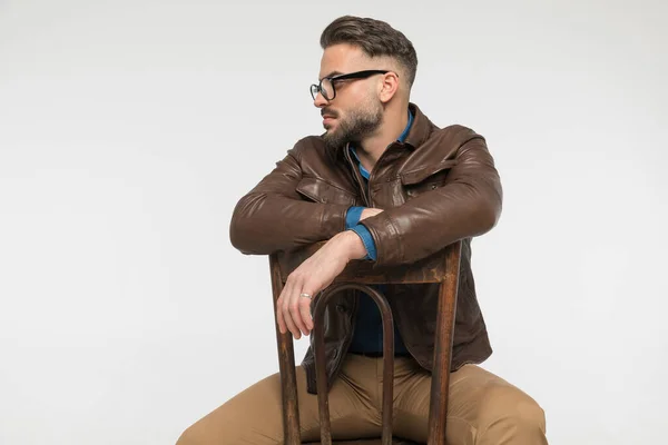 casual man in brown leather jacket holding arms on chair and looking to side while posing in front of grey background in studio