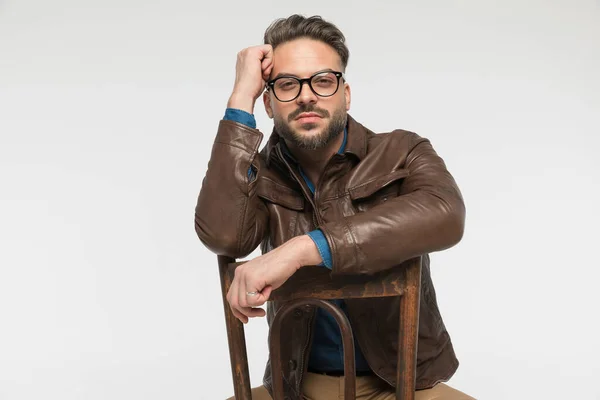 cool unshaved man in brown leather jacket with glasses holding arms and elbows on chair while sitting and posing on grey background