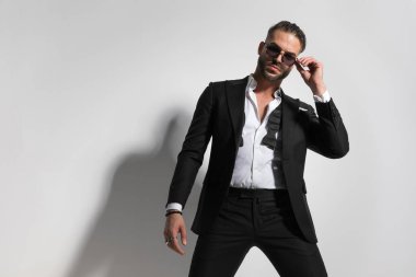 Çekici iş adamı güneş gözlüğünü takıyor ve siyah bir smokin giyiyor ve gri stüdyo arka planında çözülmemiş papyon takıyor.