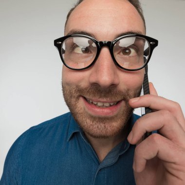 Genç adam telefonda konuşurken gözlerini şaşı yapıyor ve gri stüdyo arka planına karşı mavi gömlek ve gözlük takıyor.