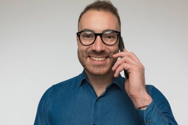 Mutlu sıradan adam telefonda konuşuyor ve gri stüdyo arka planına karşı mavi gömlek ve gözlük takıyor.