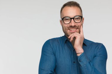 Genç sıradan bir adam kameraya gülümsüyor ve gri stüdyo arka planında mavi bir gömlek ve gözlük takıyor.