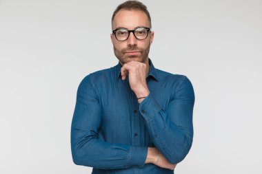 Yakışıklı, sıradan bir adam, gri stüdyo arka planına karşı mavi bir gömlek ve gözlük takarak çenesine dokunuyor.