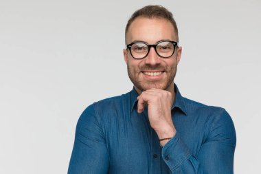 Yakışıklı sıradan bir adam tek eliyle çenesine dokunuyor ve gri stüdyo arka planında mavi bir gömlek ve gözlük takıyor.