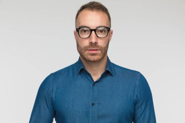 Genç adam korku içinde şaşırmış ve gri stüdyo geçmişine karşı mavi bir gömlek ve gözlük takıyor.