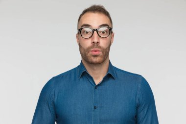 Yakışıklı, sıradan bir adam, gri stüdyo arka planında mavi bir gömlek ve gözlük takıyor.