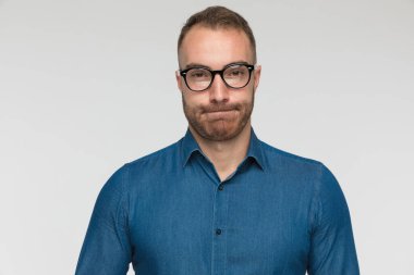 Genç adam yanaklarını şişiriyor ve gri stüdyo arka planında mavi gömlek ve gözlük takıyor.