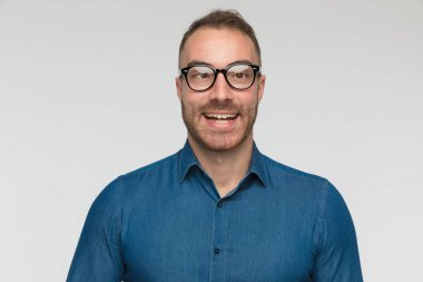 Genç adam gözlerini şaşı yapıyor, gülümsüyor ve gri stüdyo arka planında mavi bir gömlek ve gözlük takıyor.