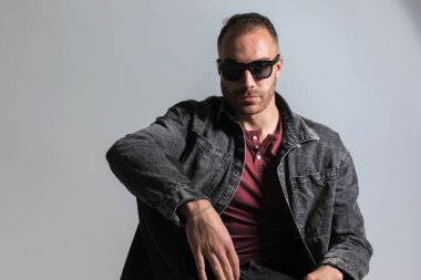 Güneş gözlüklü, dirseğini dizine dayamış ve stüdyoda gri arka planda poz veren genç bir adam.