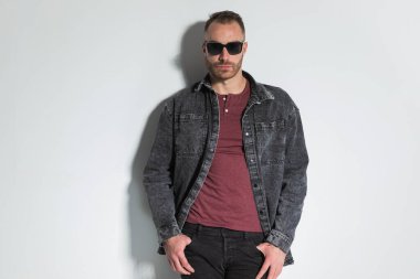 Güneş gözlüklü yakışıklı bir adamın portresi. Parmaklarını cebine sokup stüdyoda dururken gri arka planda poz veriyor.