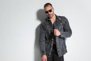 Seksi sakallı adamın portresi kot ceketi çekiyor ve ayarlıyor. Stüdyoda gri arka planda poz veriyor.